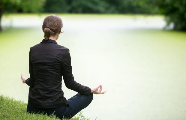 Young attractive woman sitting cross legged on lakeside, meditating, practicing yoga Easy Pose, Sukhasana, asana for meditation, pranayama, breathing, back view, copy space