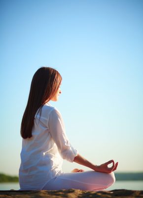 Side view of meditating woman sitting in pose of lotus against blue sky outdoors